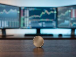 Oracle: Strong Results That Have Raised Concerns Coin standing on a wooden desk with three monitors in the background showing stock market charts.