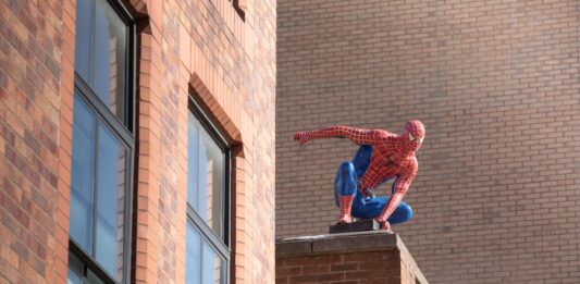 New Spider-Man Breaks North American Opening Weekend Box Office Record Spider-Man action figure crouching on a brick ledge of a building behind tall windows.