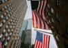Two American flags hanging between tall office buildings in a city street, viewed from below.