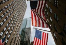 Two American flags hanging between tall office buildings in a city street, viewed from below.