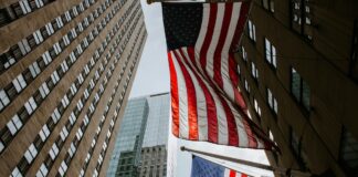 Two American flags hanging between tall office buildings in a city street, viewed from below.