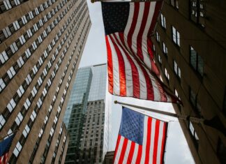 Two American flags hanging between tall office buildings in a city street, viewed from below.
