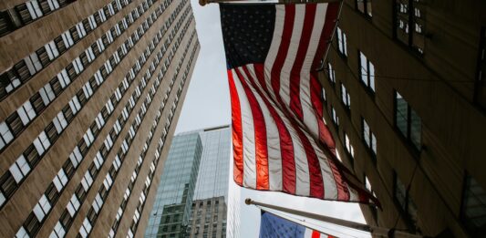 Two American flags hanging between tall office buildings in a city street, viewed from below.