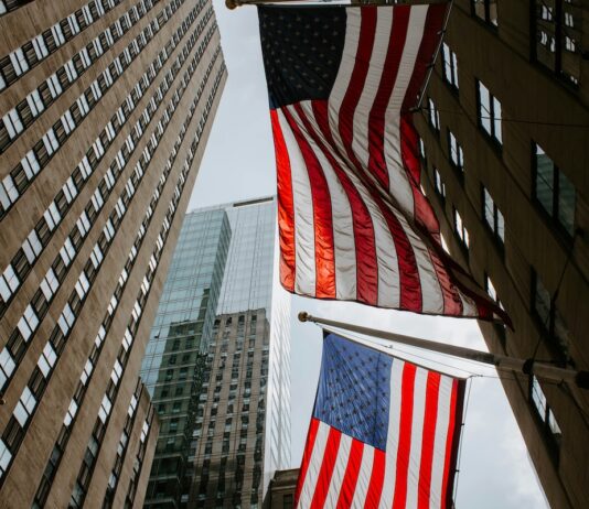 Two American flags hanging between tall office buildings in a city street, viewed from below.