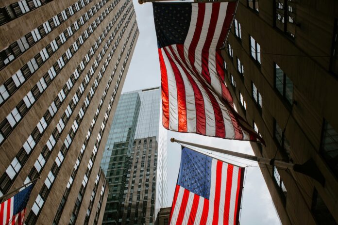 Two American flags hanging between tall office buildings in a city street, viewed from below.